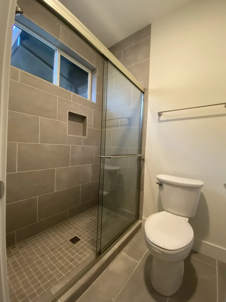 Tiled walk-in shower built by Panda Plumbing with large-format greige tile, a mosaic pan, and a sliding glass door