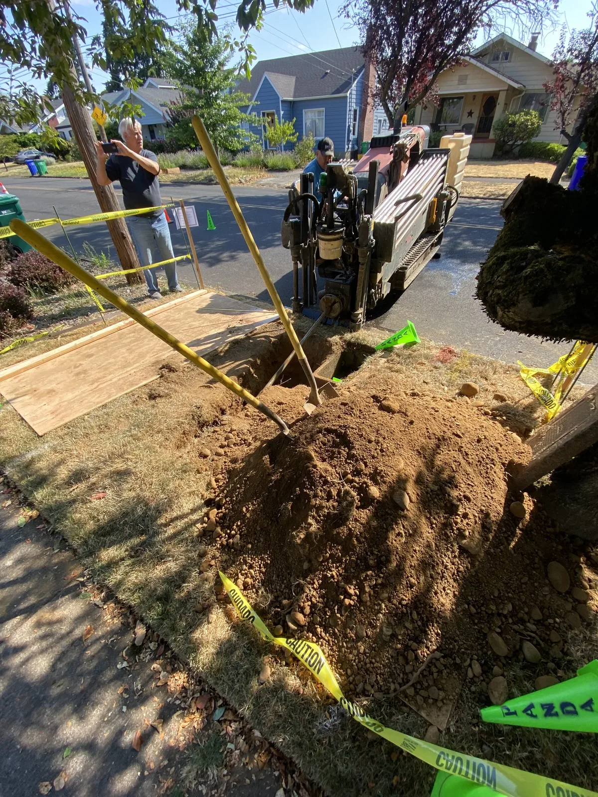 Bore pit with excavated soil and shovels, plywood protecting the lawn, and the drill rig at the street
