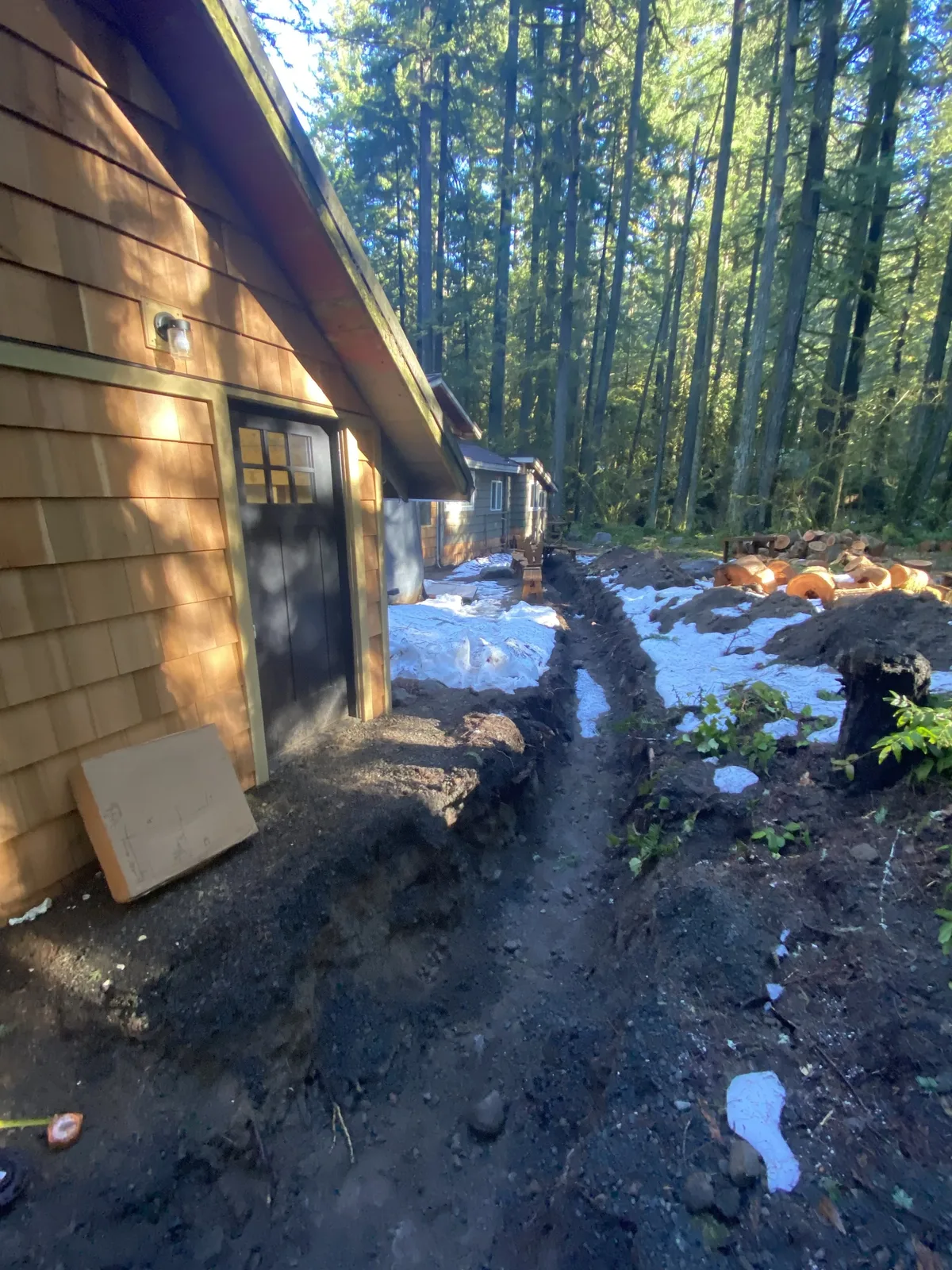 New water line trench running alongside a cedar-shingled cabin in the woods